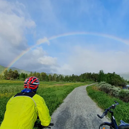 En regnbue over en grusveg med grønne dalsider på begge sidene, en syklist i forkant med rød hjelm og gul jakke. Og en sykkel med bagasje ved siden.