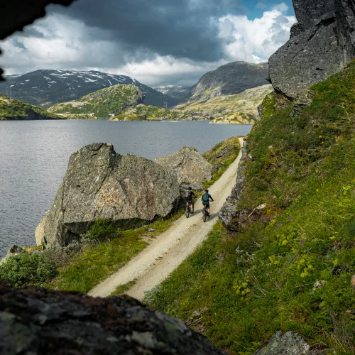 Rallarvegen langs et vann, med to syklister på vegen. Bilde er tatt gjennom et sprekk en stein, og man ser fortsatt litt snø på de mange fjelltoppene i bakgrunnen.